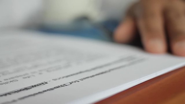A male hand edits a printed document with a pen on a wooden table.