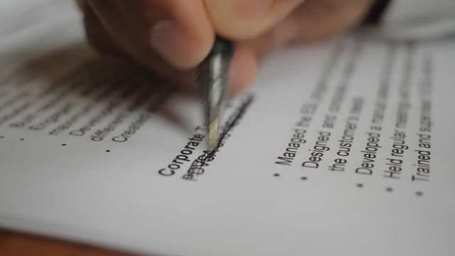 A male hand edits a printed document with a pen on a wooden table.