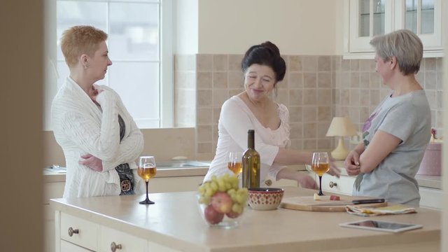 Three mature women communicating at home standing near modern table in the kitchen. Lady telling story emotionaly, actively gesticulating. Senior ladies having fun drinking wine. Camera moves right