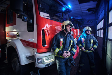 Two firemen in the fire station garage, standing next to a fire truck and looking sideways