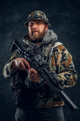 Bearded special forces soldier in the military camouflaged uniform holding an assault rifle. Studio photo against a dark textured wall