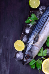 Fresh mackerel on a black chopping board surrounded by ice, lime, lemon, parsley and spices