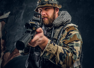 Fototapeta premium Brutal man in the military camouflaged uniform holding an assault rifle with a laser sight and aims at the target. Studio photo against a dark textured wall