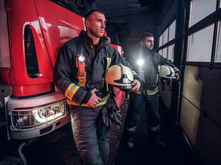 Two firemen in the fire station garage, standing next to a fire truck and looking sideways