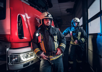 Two firemen wearing protective uniform standing next to a fire engine in a garage of a fire...