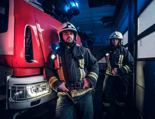 Two firemen wearing protective uniform standing next to a fire engine in a garage of a fire...