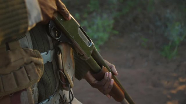 Close up following shot of an anti-poaching camouflaged ranger's gun as he walks into the early morning sunlight