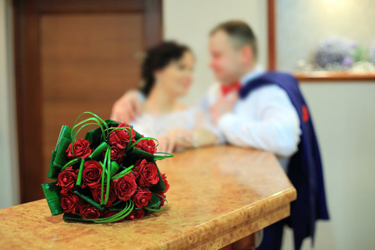 Bukiet ślubny z czerwonych r&oacute;ż i młoda para w tle.