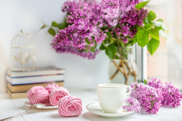 Obraz na płótnie Canvas Bouquet of lilacs, cup of coffee, homemade marshmallow and stack of books on window sill Romantic spring morning.