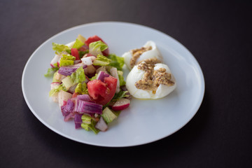Village salad (tomato; cucumber; lettuce; radish; red onion) next to two Soft-Boiled Eggs; with ground mustard on white dish on black surface. Healthy breakfast concept.