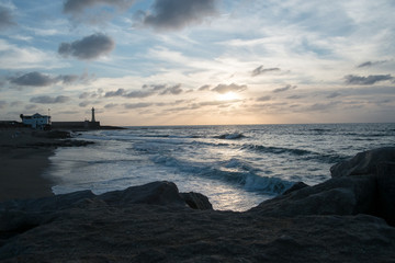 Colorful sunset in a calm ocean bay with a lighthouse and stones in the foreground.