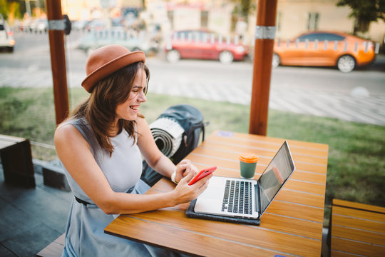 theme tourism and travel young student. beautiful young Caucasian girl in dress and hat sits in street cafe at wooden table using laptop technology and mobile phone. Search and book hotel and ticket
