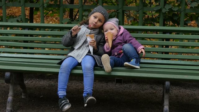 Little kids eating ice cream sitting in the park. Two sisters sitting on the bench and eating ice-cream. Funny children walking  