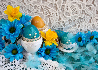 Wielkanocna kompozycja z niebieskimi pisankami, żółtymi i niebieskimi kwiatami oraz piórami na tle białej koronki © Monika