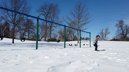 Kid walking in the playground, swing. Cute little boy (toddler, two year old) in hat with fur trim and earflaps. Child, winter time, cold weather, outdoor