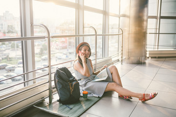 theme of tourism and travel of young student. Beautiful young caucasian girl in dress and hat sits on floor tourist rug inside terminal airport terminal. Waiting room delayed flight, delay departure