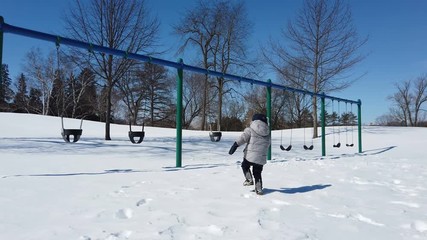 Kid walking in the playground, swing. Cute little boy (toddler, two year old) in hat with fur trim and earflaps. Child, winter time, cold weather, outdoor
