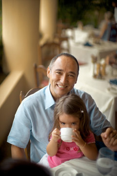 A small girl enjoys the drink as she is seated on her fathers lap.