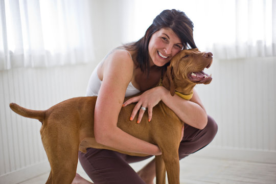Portrait of a smiling mid-adult woman hugging her dog inside a bare room in her home.