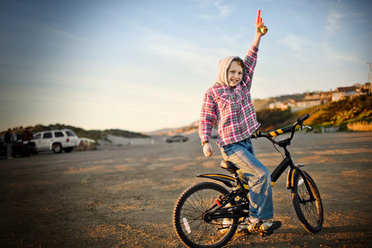 Portrait of a smiling boy in a hooded shirt pointing to the sky while straddling a bicycle in a carpark near the beach.