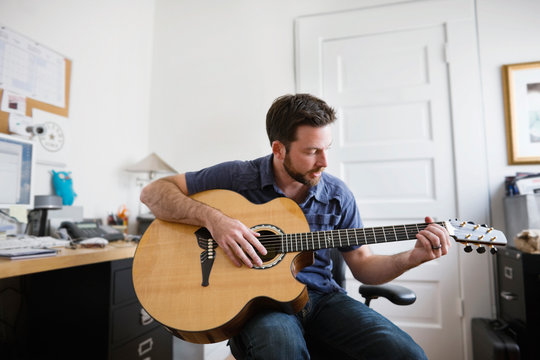 Young adult man playing an acoustic guitar in a home office.