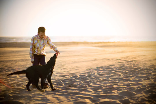 Boy playing with his black dog on a sandy beach at sunset.