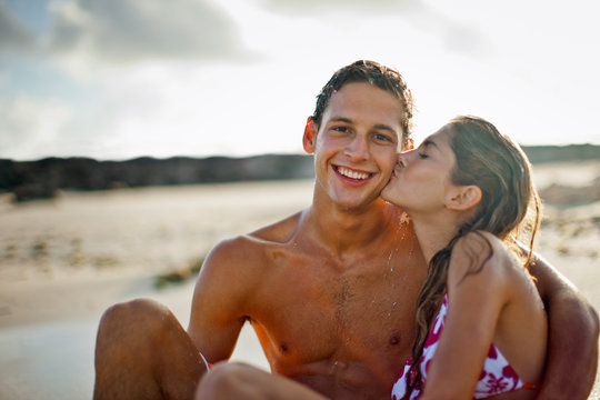Portrait of a smiling young man being kissed on the cheek by his girlfriend on a sandy beach.