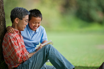 Happy father and son reading a book under a tree.