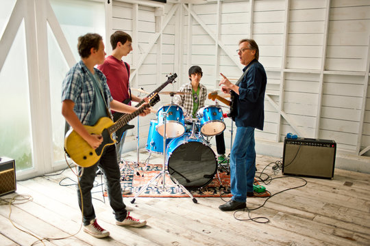 Mid adult man talking to three teenage boys playing with musical instruments inside a garage.
