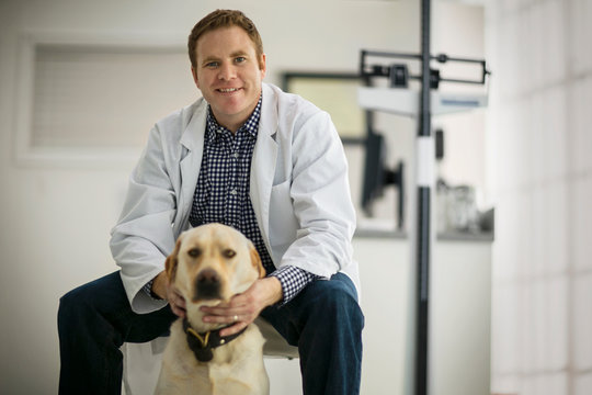 Portrait of a smiling male doctor patting a dog inside an exam room.