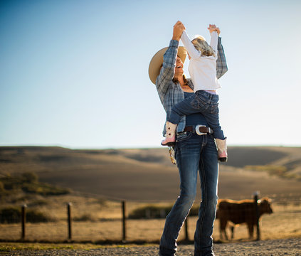 Farmer's wife joyfully lifting her toddler into the air while out on the ranch.
