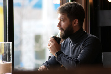 handsome bearded guy drinks a drink from a straw near the window in a cafe.  attractive young stylish man sitting in a cafe and looking out the window. Business, freelance