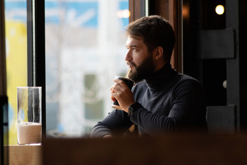 handsome bearded guy drinks a drink from a straw near the window in a cafe.  attractive young stylish man sitting in a cafe and looking out the window. Business, freelance