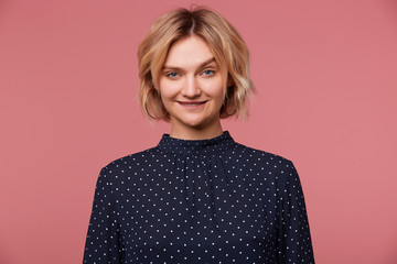 Close up of playful young beautiful attractive blonde woman dressed in blouse with polka dots, coquets, flirting, biting lip shows attraction, isolated over pink background