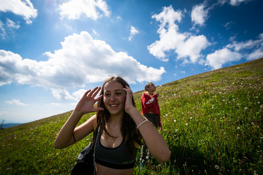 Two happy young girls are on the slope of a blooming hill in the Italian Alps. The first smiles, adjusting her hair and the second blows on a flower. Green meadow, blue sky and sun with white clouds
