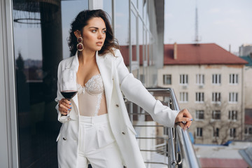 Young beautiful sexy woman in white corset and jacket relaxing at balcony with a view to the urban landscapes and drinking red wine.