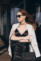 Beautiful young sexy woman in sunglasses, glamour girl in the white elegant jacket, black dress, makeup pastel tone lipstick, nails on the office balcony with the urban background.