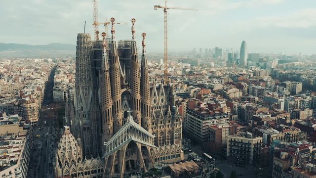 Aerial; drone view of main Barcelona attraction; majestic building towering over the rooftops of Eixample; sharp domes and unusual forms of great unfinished work of neo-gothic art; camera moves around