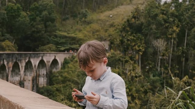 Medium shot happy cute little European boy taking smartphone photo at Ella Nine Arches Bridge on Sri Lanka vacation.