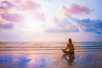 Fototapeta na wymiar Young beautiful woman sitting on sunset sea beach and practicing zen yoga in lotus pose. Evening meditation with view on surrealistic sky and ocean. Reflection on wet sand