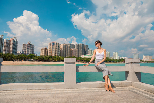 Cheerful and beautiful girl resting fun in vacation, on the pier on background of city. In China, Hainan city of Sanya. View of island in bay. Town Sanya is popular tourist destination in China