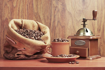 A cup filled with grains of coffee, a coffee grinder, a bag of coffee on a wooden table.
