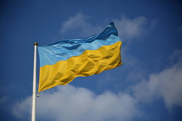 National flag of Ukraine, big, hangs on white pole, waves on wind, in background blue sky with nice white clouds