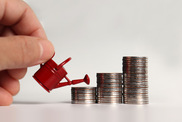 A hand holding a watering can and three piles of stepped coins. © Hyejin Kang