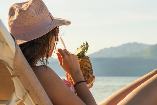 Young beautiful woman with hat drinks pina colada while sunbathing on the beach in vacation. 