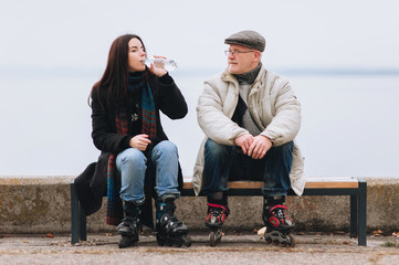 A girl on rollers drinks water from a bottle. Sport and water. Father and daughter on roller skates sit on a bench. Active old people.