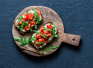 Tomatoes and rocket salad whole grain bread bruschetta on a wooden chopping board on a dark background, top view. Copy space