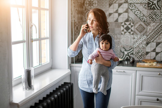 busy serious mother holding her baby and talking on the smartphone in the kitchen. close up photo, free , spare time