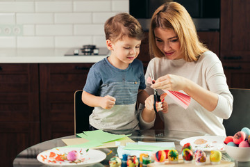 It is so funny to prepare to Holy Easter with children. Attractive blonde woman is teaching her adorable little kid how to cut out Easter decor from the coloured paper , sitting at kitchen
