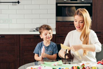 It is so funny to prepare to Holy Easter with children. Attractive blonde woman is teaching her adorable little kid how to cut out Easter decor from the coloured paper , sitting at kitchen
